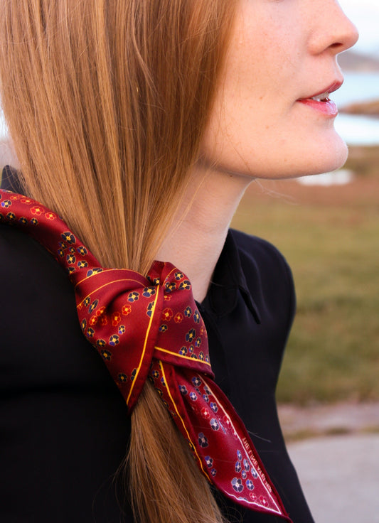 Close-up of a woman's hair with Hearts Ruby silk scarf by The Line Atelier, diamond size, with gem toned pansy flowers on a blurred Scandinavian coast background