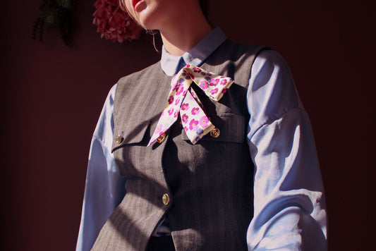 Person wearing a gray vest over a blue shirt with the pink floral scarf Hearts Ease silk twilly by The Line Atelier around the neck against a dark background