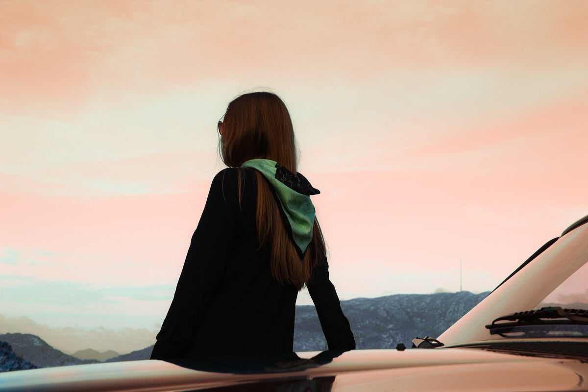 Person standing by a car with a scenic view in the background wearing Ballet - The Poised Rogue 60x60cm cool green silk scarf with black ballerina lineart