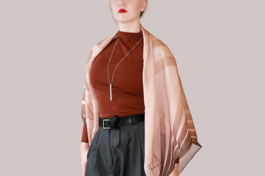 A woman wearing a blue and light blue striped shawl, Bronze Sandrift silk sand wool by The Line Atelier, draped over her shoulders, styled with a rust-brown mock-neck top, a black belt, and dark gray trousers, set against a solid beige background.