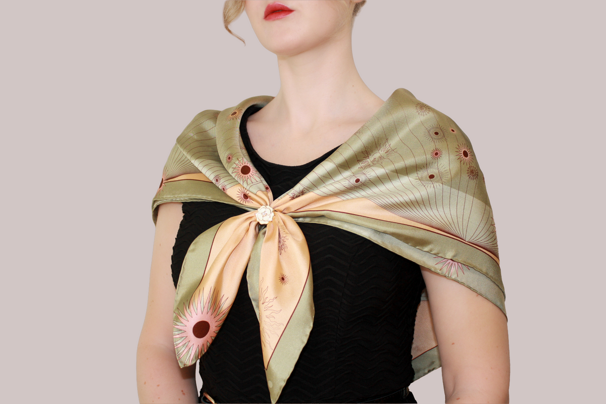 A woman wearing a silk scarf, Courage of Leo by The Line Atelier, tied around her shoulders, featuring a light green and peach Leo constellation pattern with small red floral accents and a decorative brooch at the center knot, set against a solid taupe background.