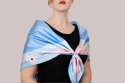 A light blue and pink celestial patterned silk scarf, Gemini Lovers by The Line Atelier, tied around a model's neck over a black top, set against a solid taupe background.