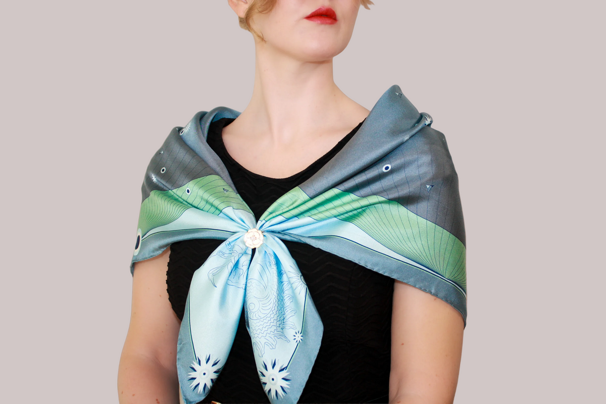 A woman wearing a blue-green Gentle Caner constellation silk scarf with white starburst patterns and green striped borders, shown against a solid taupe background.