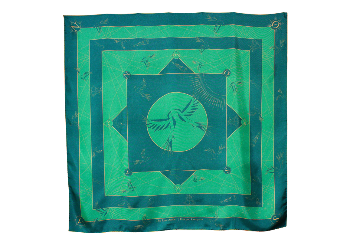 A large square silk scarf, Halcyon Compass by The Line Atelier, with a green and teal geometric pattern featuring a central bird motif, hanging on a brown wall.
