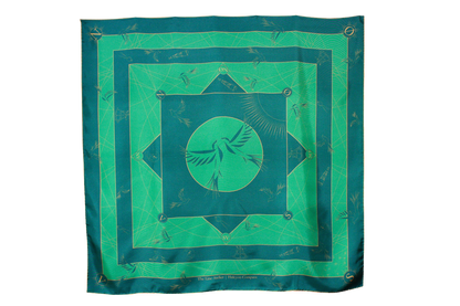 A large square silk scarf, Halcyon Compass by The Line Atelier, with a green and teal geometric pattern featuring a central bird motif, hanging on a brown wall.