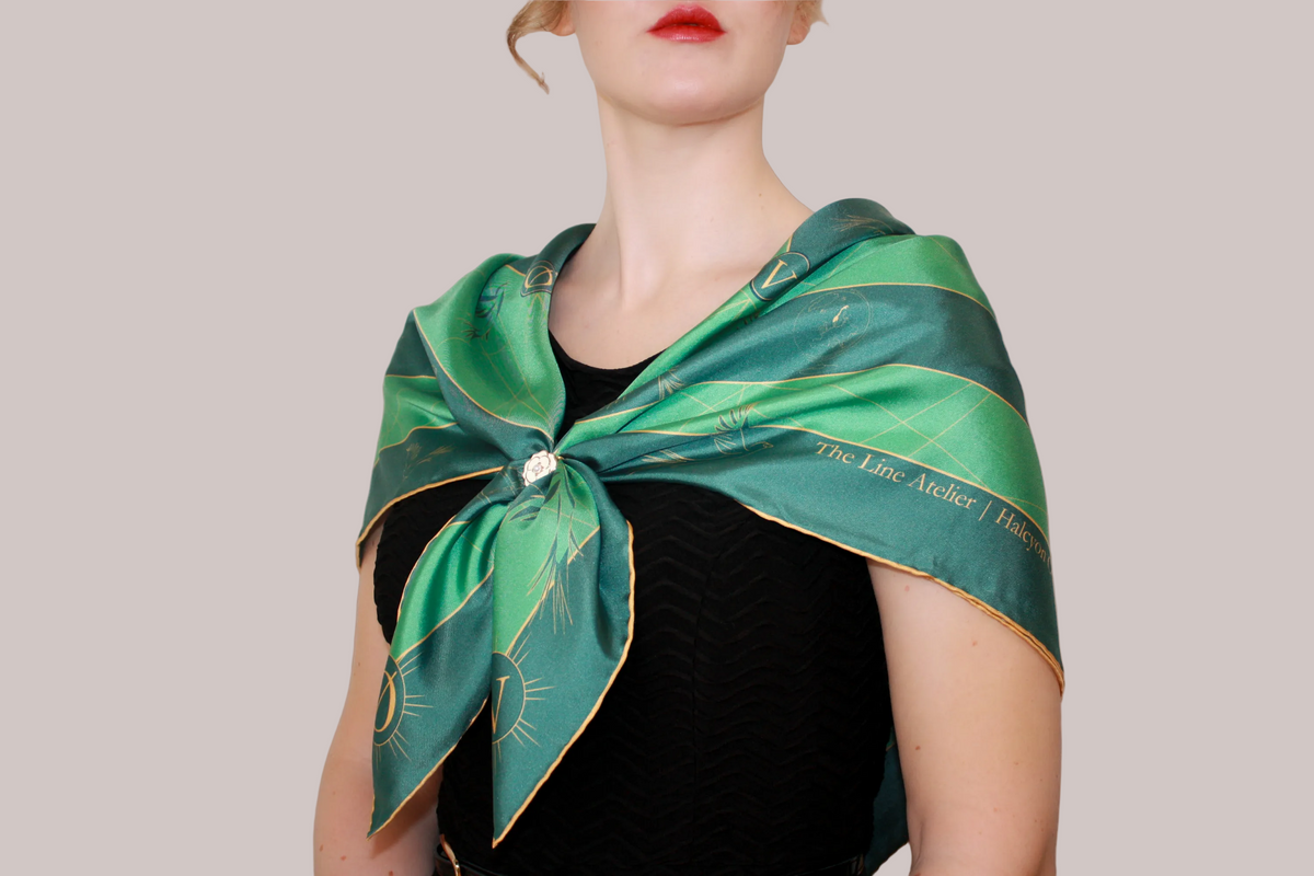 A woman wearing a green silk scarf, Halcyon Compass by The Line Atelier, with gold celestial patterns and text, tied around her neck over a black top, against a solid tan background.