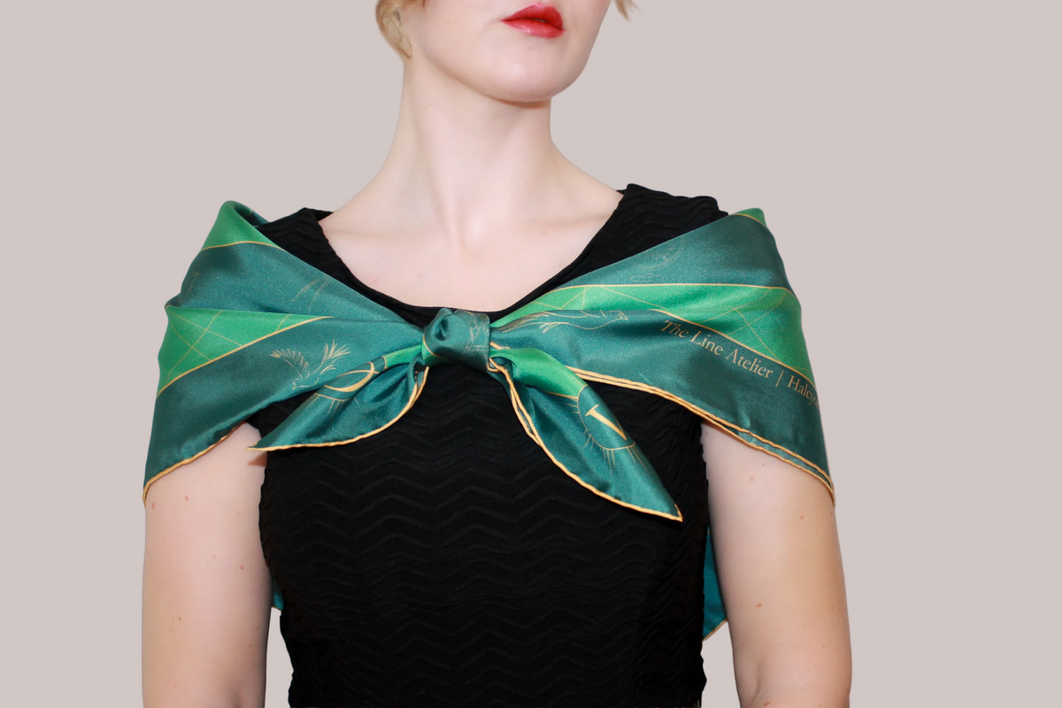 A woman wearing a green silk scarf, Halcyon Compass by The Line Atelier, with gold celestial patterns and text, tied around her neck over a black top, against a solid tan background.