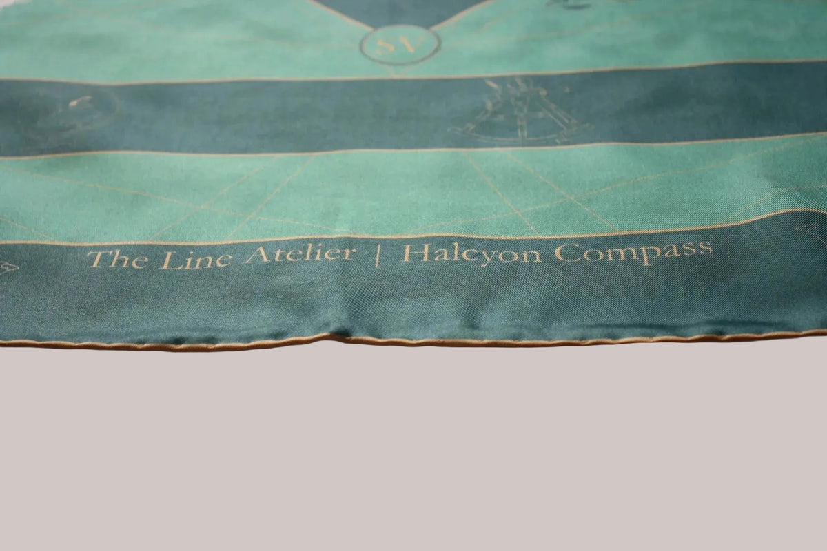 A close-up of a teal and dark teal silk scarf with gold embroidery, featuring the text 'The Line Atelier | Halcyon Compass' and a nautical compass design, laid over a white fabric background.