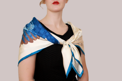 A close-up of a model wearing a cream-colored silk scarf Halcyon Seas with a blue and brown feather pattern, styled over her shoulders.