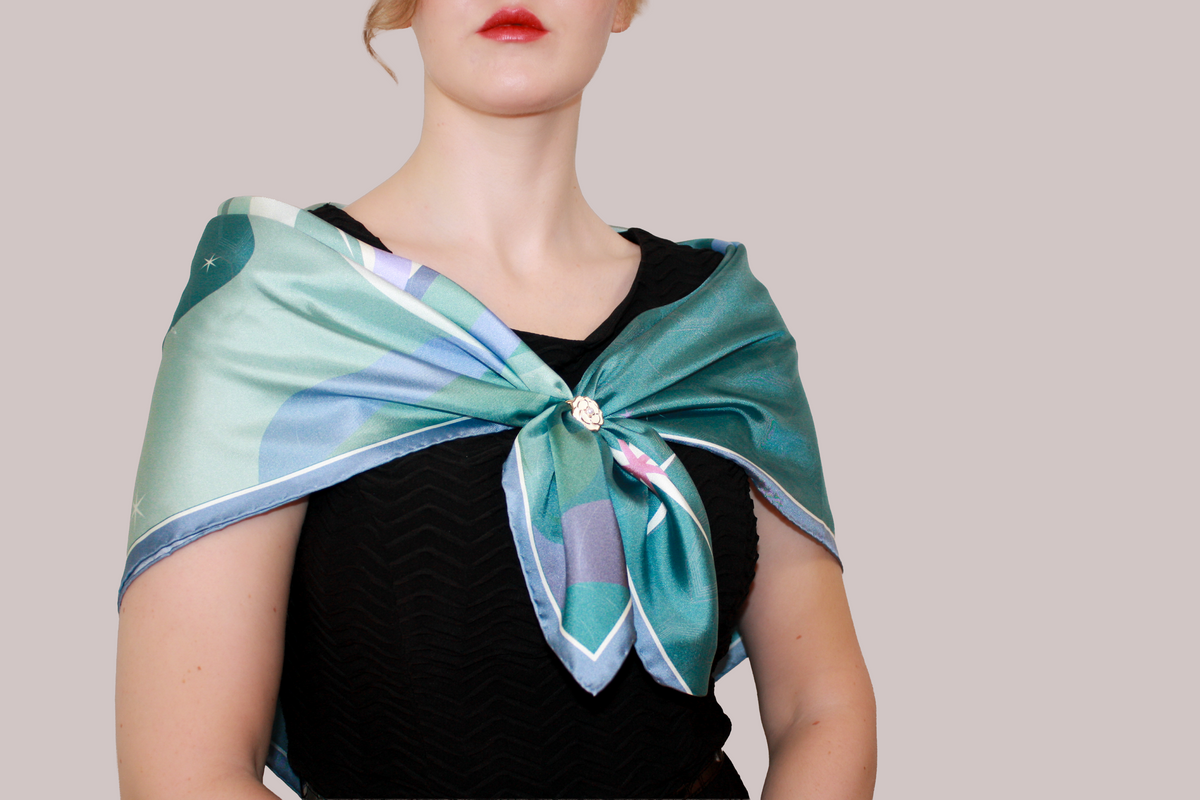 A close-up of a woman wearing a light blue and mint green striped silk scarf, Halcyon Medusa by The Line Atelier, tied around her neck, paired with a black chevron-patterned top, set against a solid grey background.