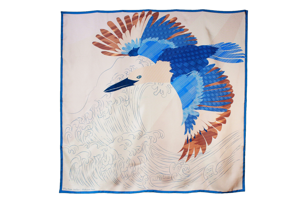 A large square silk scarf, Halcyon Seas by The Line Atelier, featuring a stylized blue and brown bird in flight over white waves, bordered in blue, displayed flat against a brown wall.