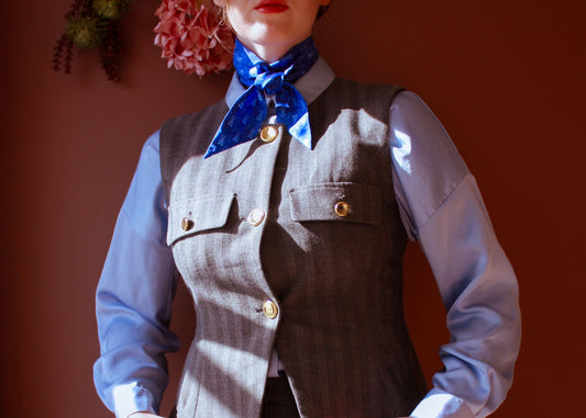 Person wearing a vest with gold buttons and the blue Ocean Rogue silk twilly scarf by The Line Atelier against a brown background