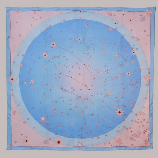 A square silk scarf featuring a celestial design of the Gemini constellation with a large light blue circle, constellation lines, and small stars on a soft pink background, finished with a light blue border and line art of ballet dancers in pairs.