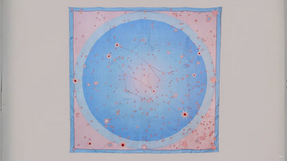 A square silk scarf featuring a celestial design of the Gemini constellation with a large light blue circle, constellation lines, and small stars on a soft pink background, finished with a light blue border and line art of ballet dancers in pairs.