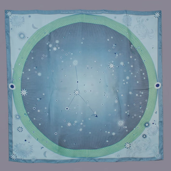 A square scarf featuring a celestial design with a light blue starry sky, a green ring, and the Cancer constellation patterns, hanging on a brown wall.