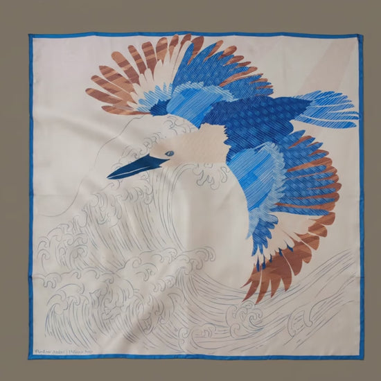 A large square silk scarf, Halcyon Seas by The Line Atelier, featuring a stylized blue and brown bird in flight over white waves, bordered in blue, displayed flat against a brown wall.