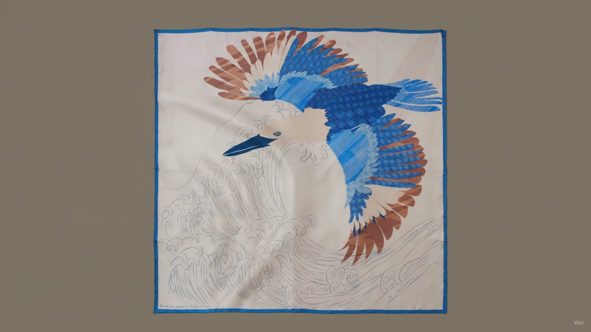 A large square silk scarf, Halcyon Seas by The Line Atelier, featuring a stylized blue and brown bird in flight over white waves, bordered in blue, displayed flat against a brown wall.