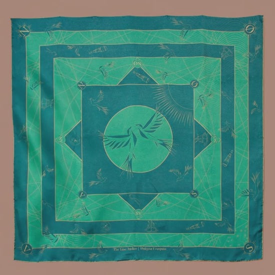 A large square silk scarf, Halcyon Compass by The Line Atelier, with a green and teal geometric pattern featuring a central bird motif, hanging on a brown wall.