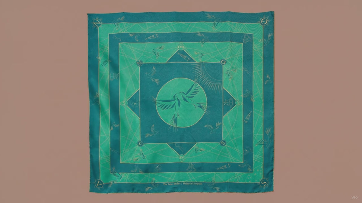 A large square silk scarf, Halcyon Compass by The Line Atelier, with a green and teal geometric pattern featuring a central bird motif, hanging on a brown wall.