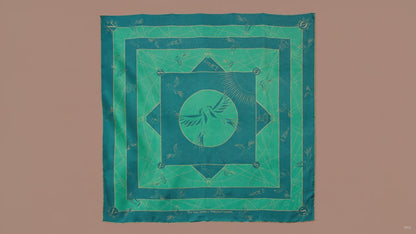 A large square silk scarf, Halcyon Compass by The Line Atelier, with a green and teal geometric pattern featuring a central bird motif, hanging on a brown wall.