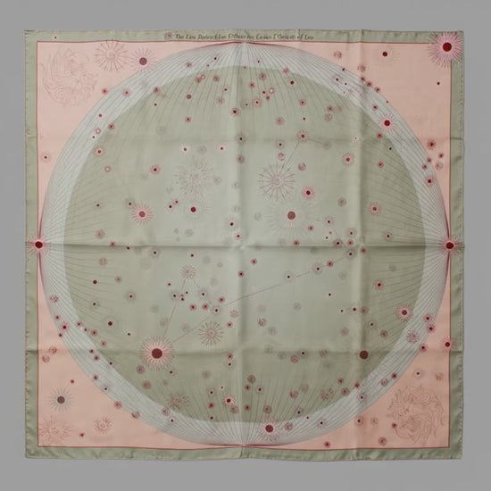 A square silk scarf featuring a celestial design with a large central pale green circle containing stars and the Leo constellation, surrounded by a peach-pink border and a muted sage green edge.