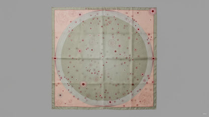 A square silk scarf featuring a celestial design with a large central pale green circle containing stars and the Leo constellation, surrounded by a peach-pink border and a muted sage green edge.